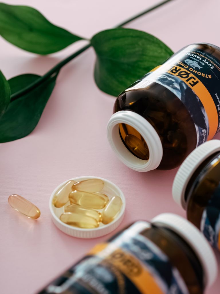Vitamin capsules spilling from bottles with green leaves on a pink surface.
