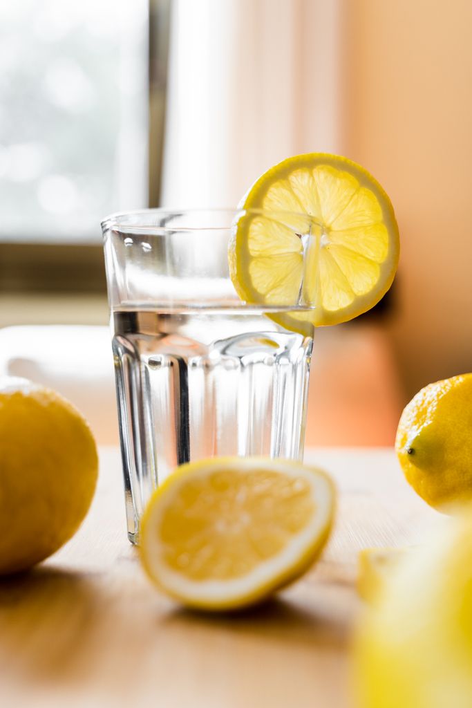 Glass of pure refreshing water served on wooden table near scattered sliced and whole lemons in kitchen in sunny morning